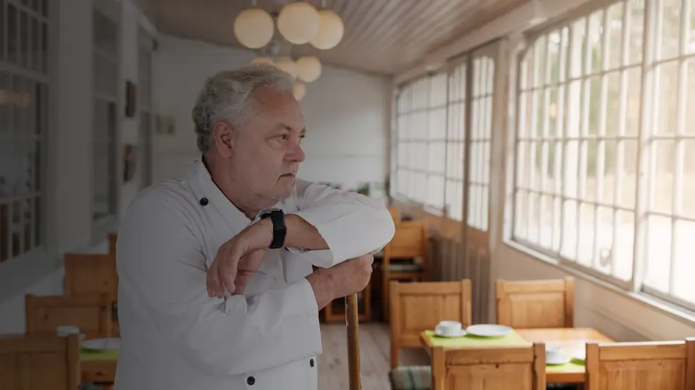 Cocinero apoyado en la barra de su restaurante vacío, observando la sala sin clientes.