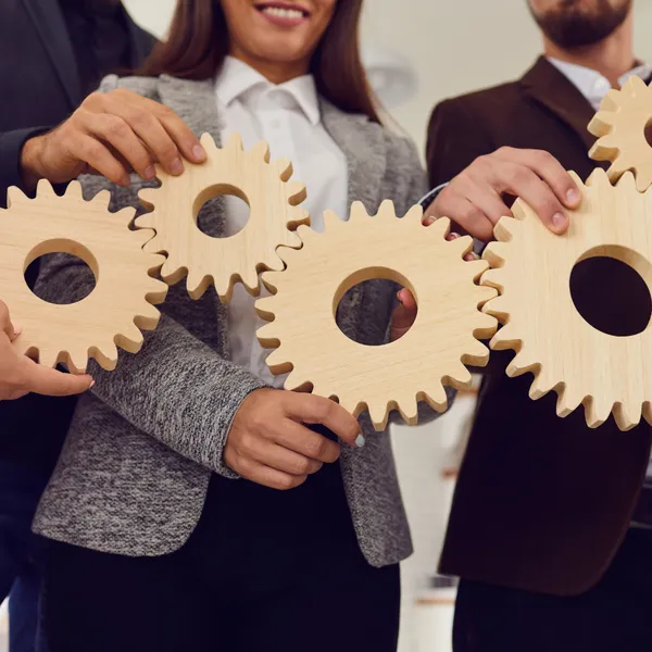Personas profesionales vestidas formalmente sujetando engranajes de madera entrelazados. Representa colaboración estratégica, coordinación entre áreas y visión conjunta.