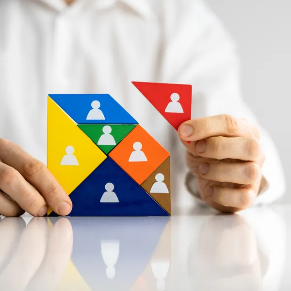 Manos construyendo una figura de equipo con bloques de colores, cada uno representando personas o roles distintos. Transmite estrategia, estructura y selección precisa.