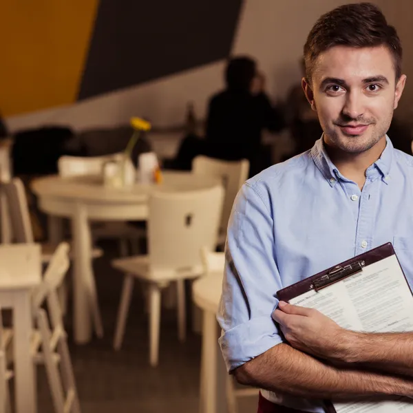 Hombre joven con camisa clara y portapapeles en la mano, en un entorno de restaurante moderno. Fondo con mesas y clientes desenfocados.
